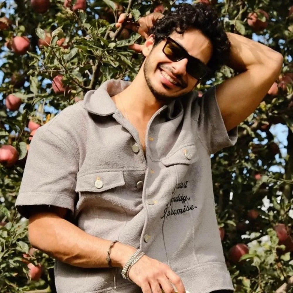 Ishaan Khatter wearing orionz jewels in a grey shirt with text standing in an apple orchard