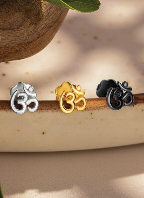Three small decorative orionz om studs in silver, gold, and black on a wooden surface with a natural background.