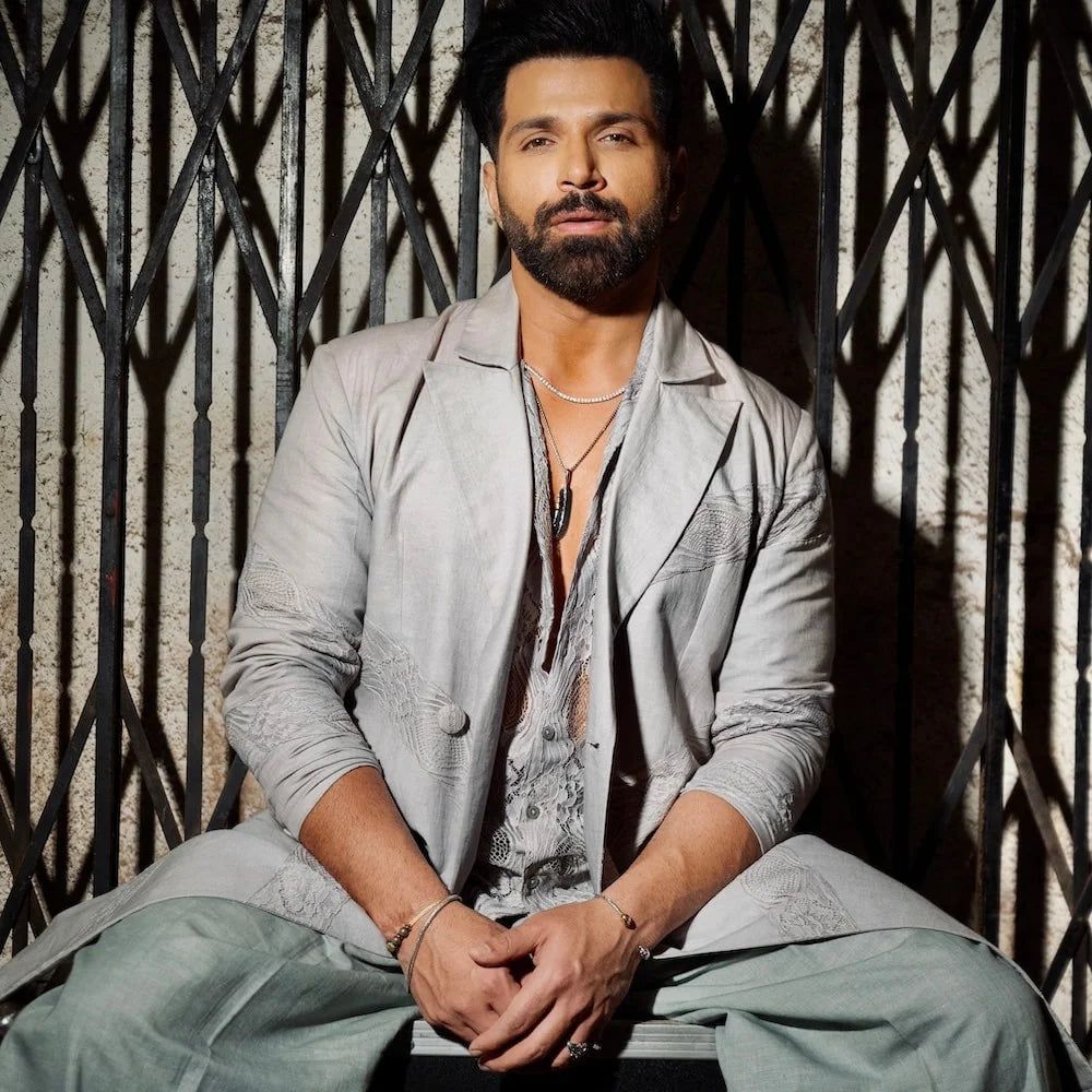Rithvik Dhanjani in a light gray suit sitting against a textured wall with geometric patterns wearing orionz jewels