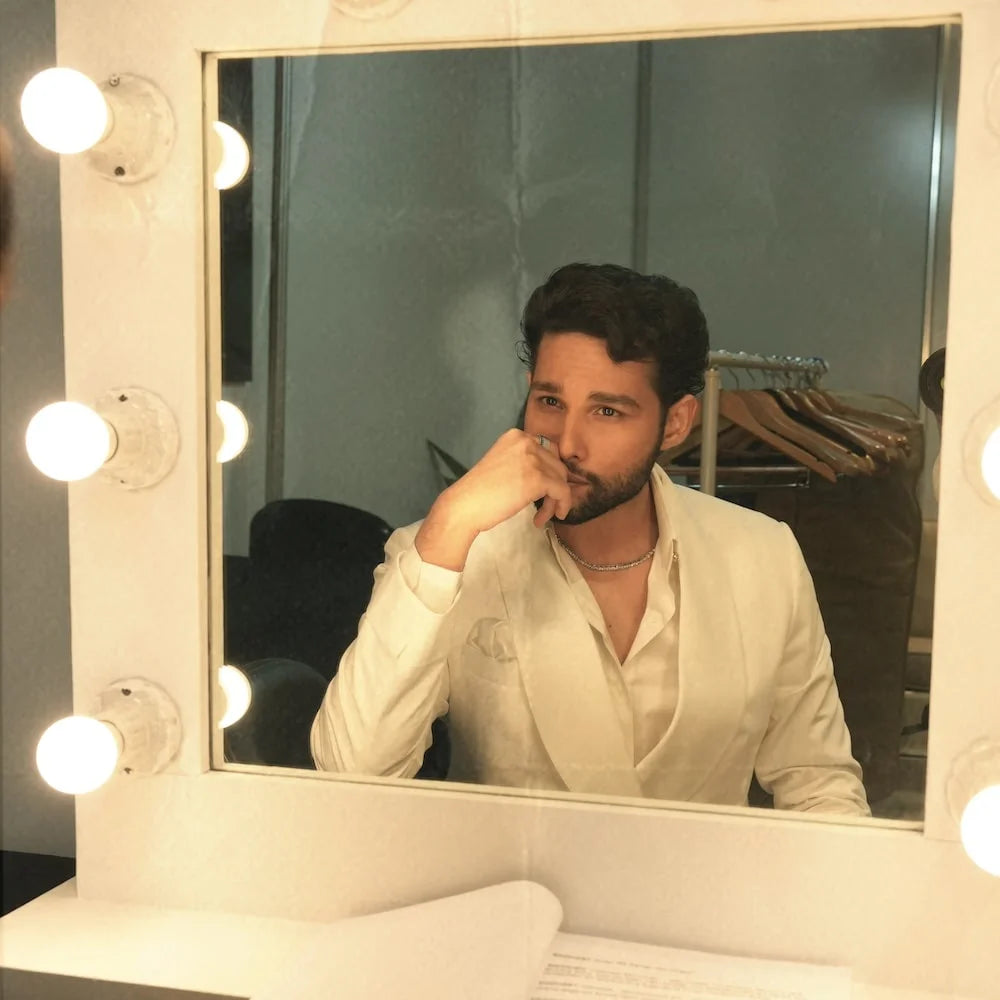 Siddhant Chaturvedi in a white shirt sitting in front of a mirror with lights, wearing a orionz jewels necklace.