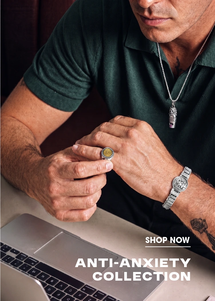 A man in a dark green polo shirt holds a small gold ring between his fingers, wearing a silver pendant necklace and a silver watch. The image features a laptop and includes text overlays reading 'SHOP NOW' and 'ANTI-ANXIETY COLLECTION'.
