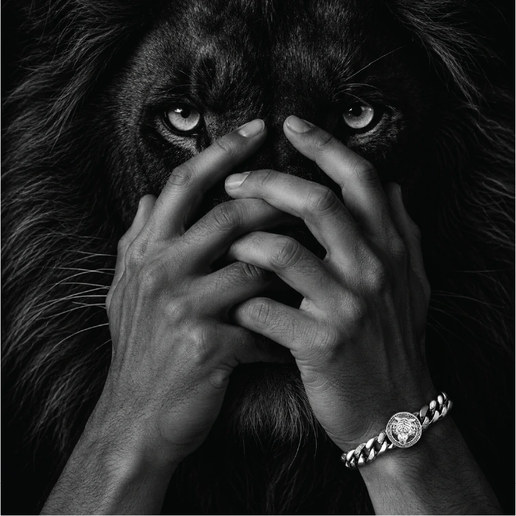 Black and white close-up of a lion face with a person's hands covering the mouth area, wearing a chunky silver chain watch with a round medallion face on the wrist.