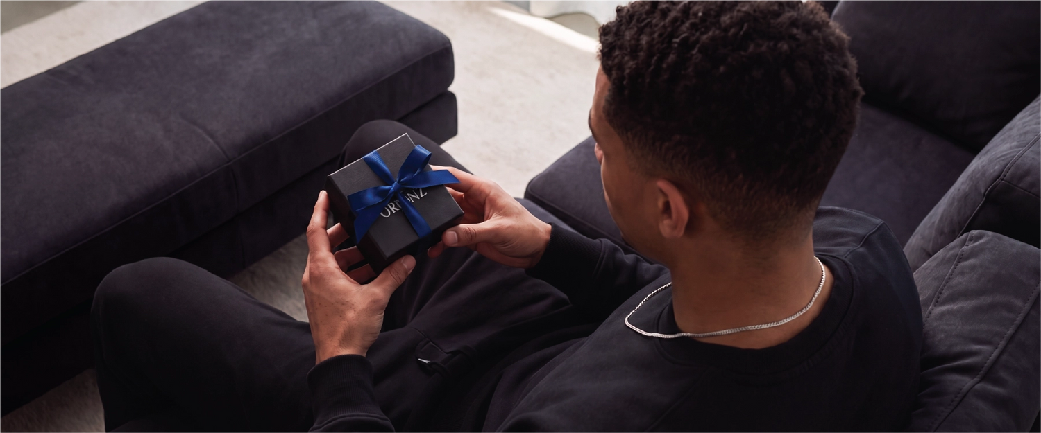 A man sitting on a dark gray couch holds a small black gift box tied with a blue ribbon. The box has the Gucci logo printed on it.