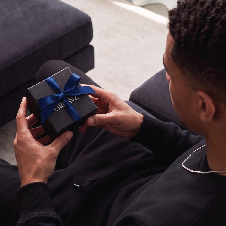 A person seated indoors holds a small black gift box tied with a blue ribbon. The box has the brand name UK PRIZ visible on it.