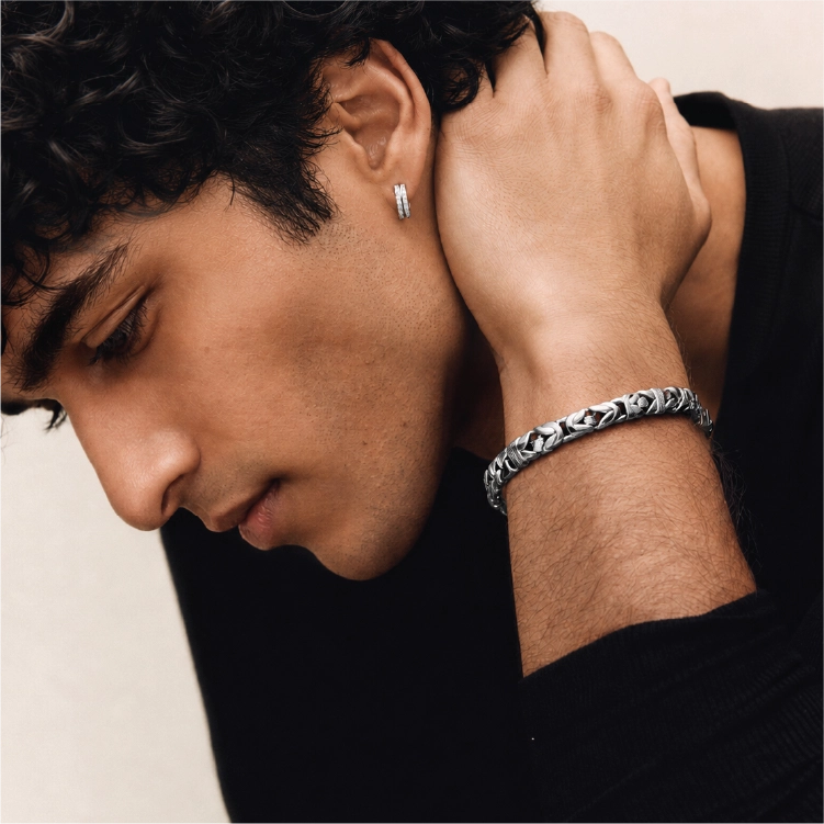 A close-up lifestyle shot of a man wearing a textured silver chain bracelet on his wrist and a matching silver hoop earring in his ear. He is wearing a black long-sleeve shirt against a neutral light beige background.