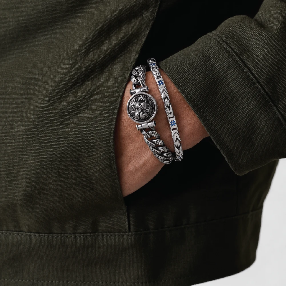 A close-up of a person's wrist wearing two silver-toned bracelets, one with a textured rope chain and the other with an engraved circular medallion and chain, set against an olive green jacket.