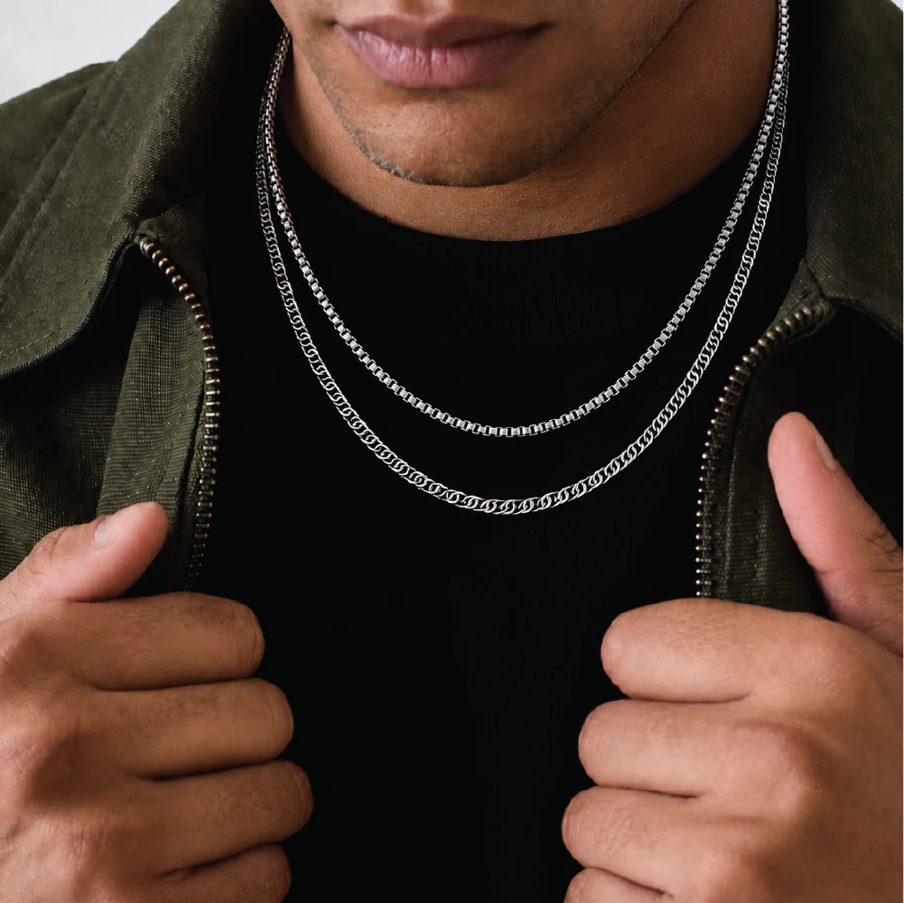 A close-up lifestyle shot of a man wearing two silver chain necklaces over a black t-shirt and an olive green zip-up jacket.