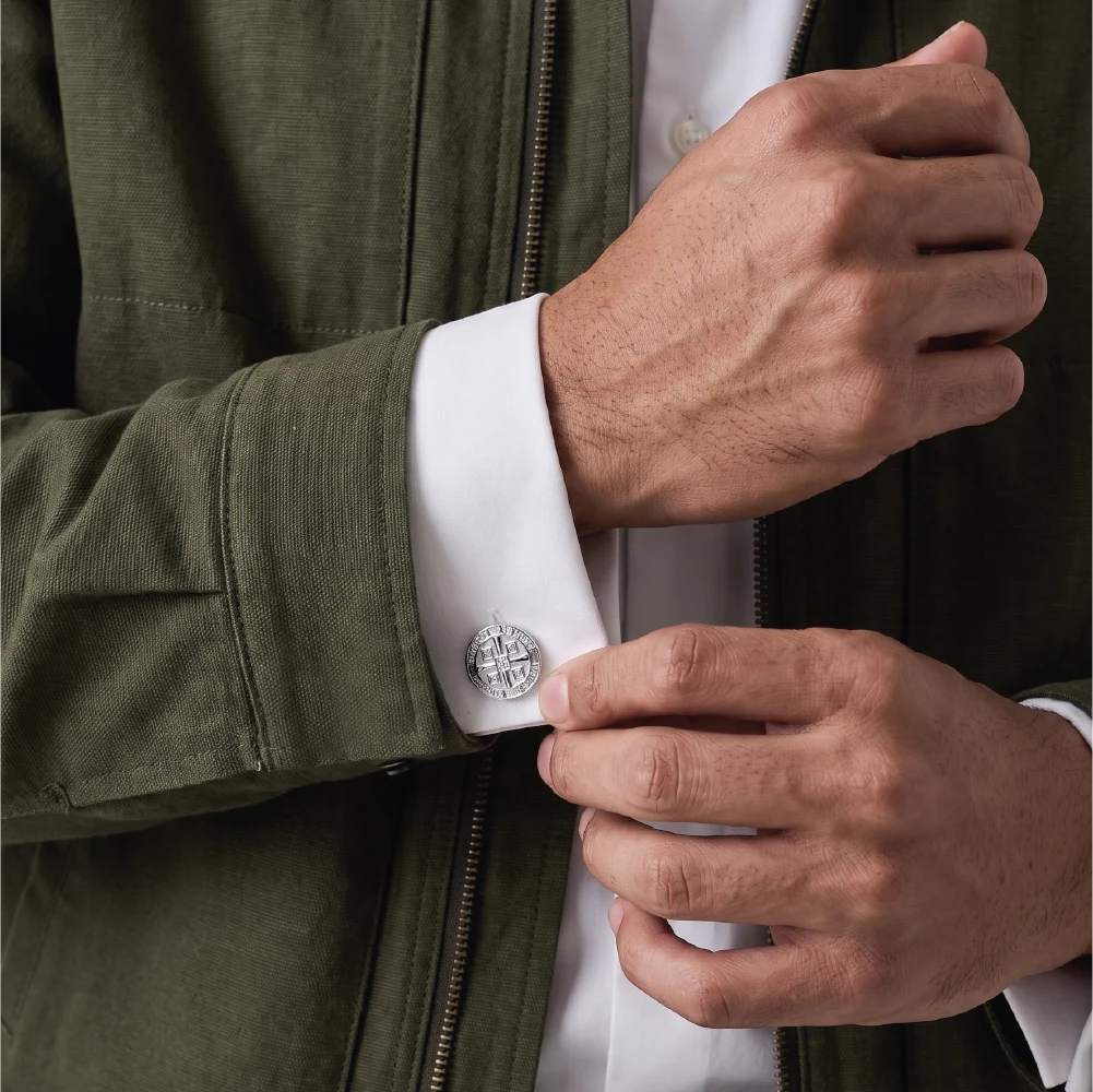 Close-up of a man's hands adjusting a silver compass cufflink on a white dress shirt cuff, wearing an olive green jacket.