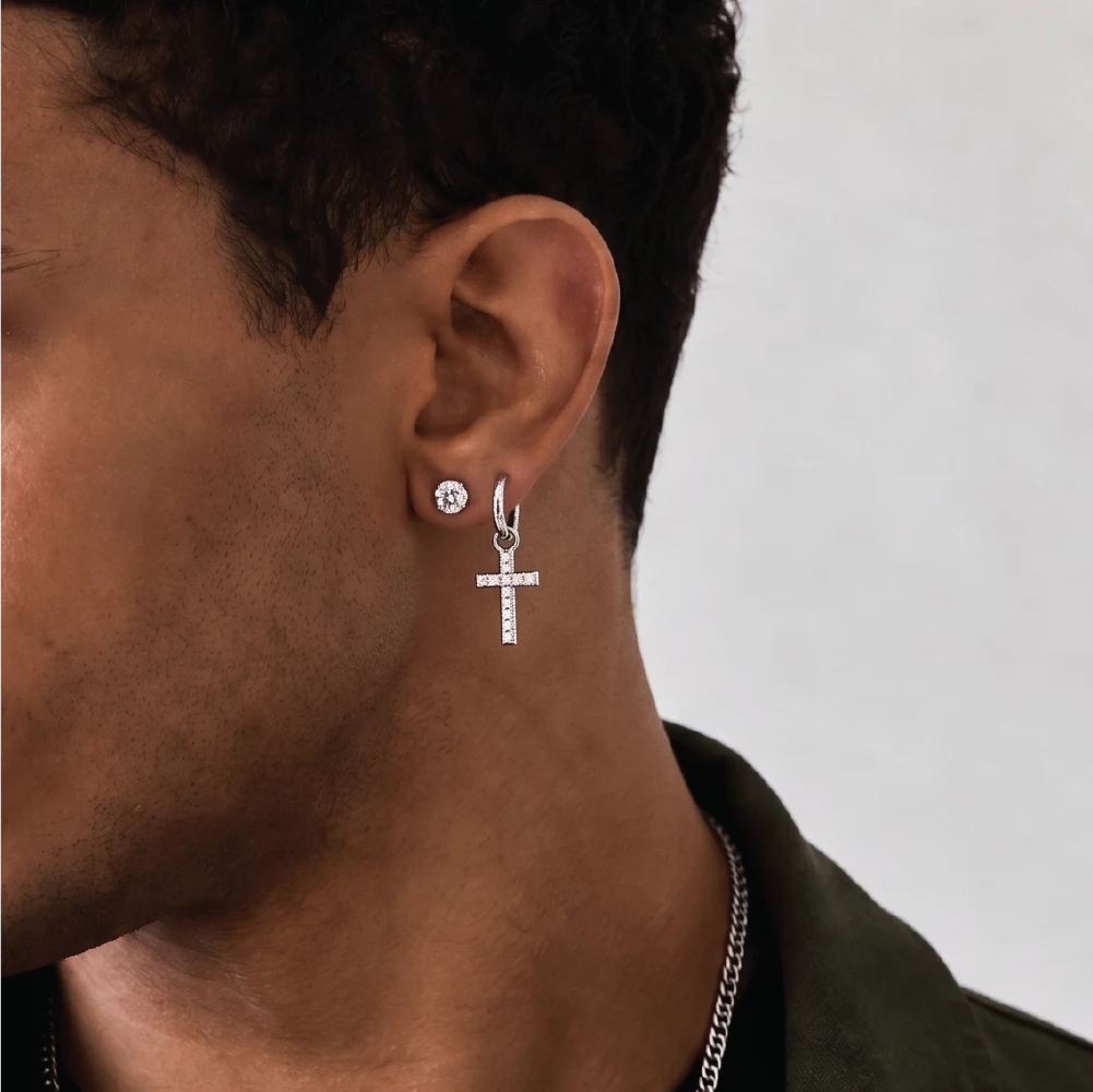 A close-up of a man's ear wearing a silver hoop earring with a dangling cross charm, set against a light gray background.
