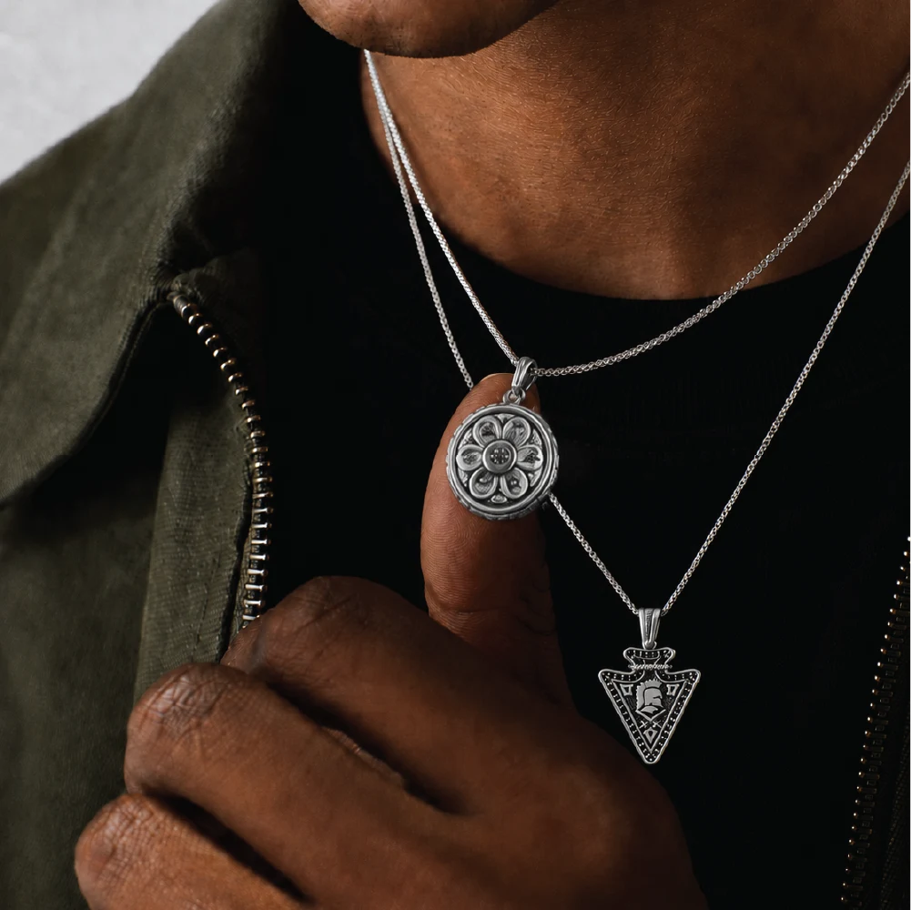 A close-up of a person wearing layered silver necklaces with ornate pendants over a black shirt and olive green jacket.