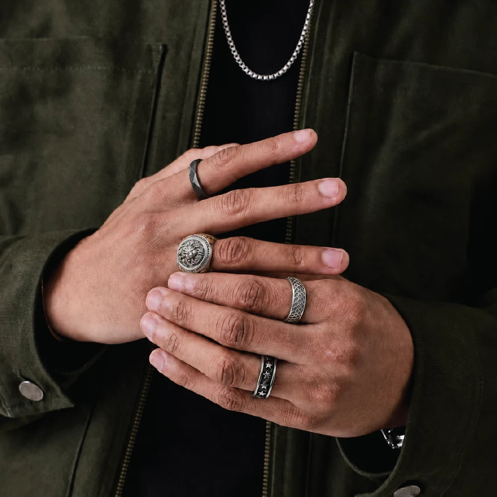 A close-up of a person's hands wearing multiple silver rings, including a large signet-style ring, a textured band, and a ring with a dark inlay, styled with an olive green jacket.