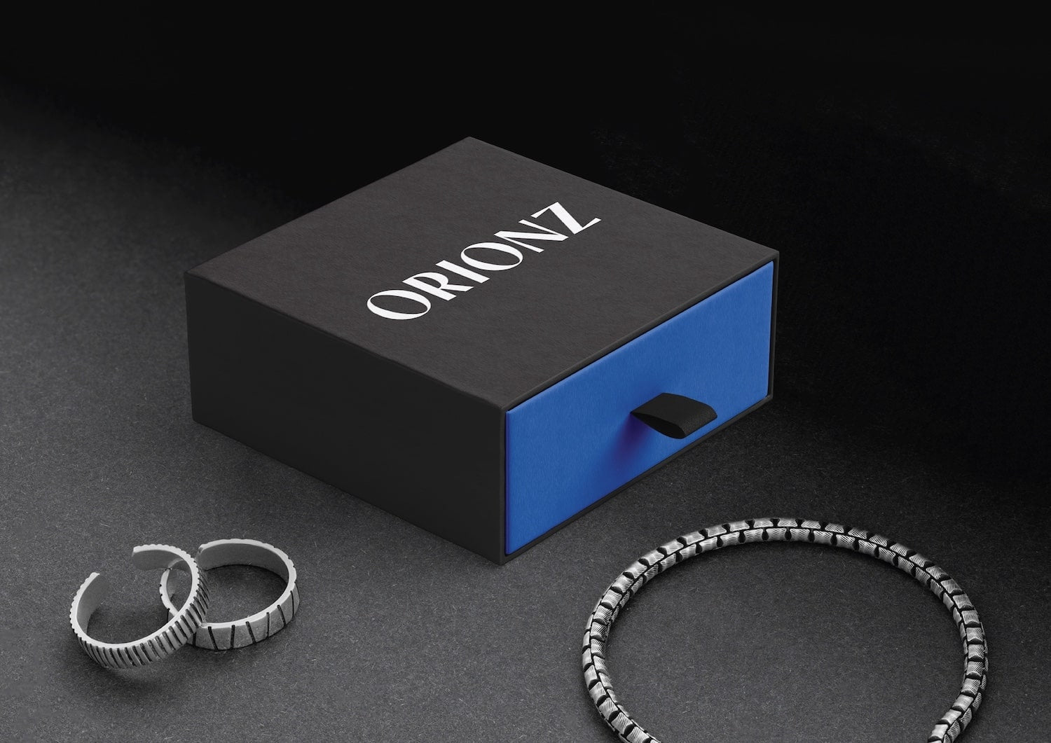 Black 'ORIONZ' jewelry box with rings and a bracelet on a dark surface