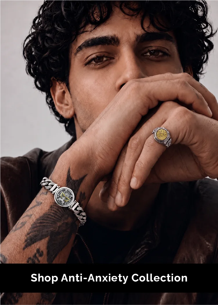 A close-up lifestyle shot of a man with curly dark hair wearing a silver-toned watch with a green dial and a matching silver ring with a yellow center. He is wearing a brown leather jacket and has a tattoo on his arm.