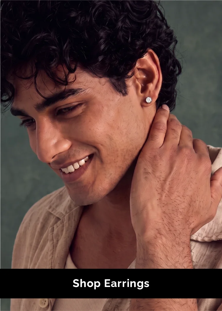 A smiling man with curly dark hair wears a small round stud earring, touching his neck. The background is a muted green textured backdrop, and a black banner at the bottom reads 'Shop Earrings'.