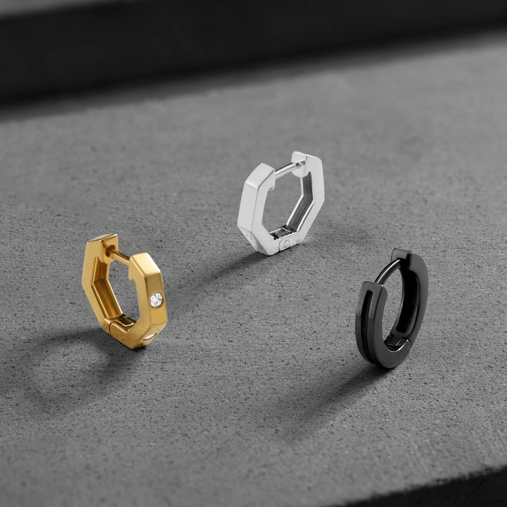 Three orionz hexagonal hoop earrings in gold, silver, and black on a gray surface.
